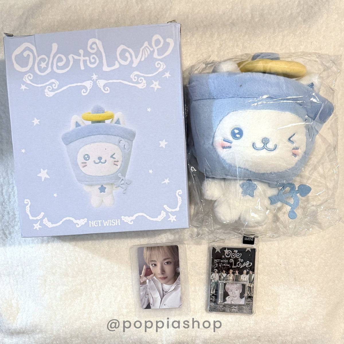 NCT WISH 'Ode to Love' plush toy set on a white textured fabric background, including the light blue illustrated packaging box, the plush in clear plastic packaging, and two photocard inserts.